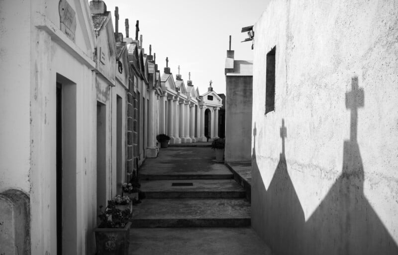 A narrow passage between rows of above-ground tombs in a cemetery, with shadows of crosses cast on the walls. The black and white image highlights the architectural details and serene atmosphere.