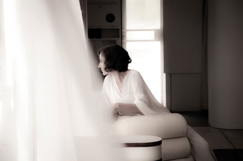 A woman in a loose, white outfit sits on a white armchair, gazing out of a window. Soft light filters through sheer curtains, creating a peaceful and contemplative mood in a minimalist setting.