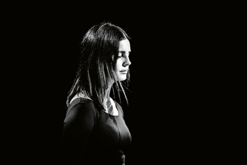 A woman with long, dark hair stands in profile against a black background, eyes closed. The lighting creates a dramatic contrast, highlighting her face and upper body while the rest blends into the dark.