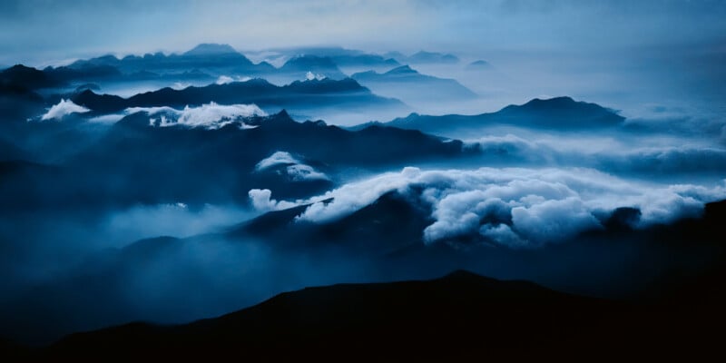 A breathtaking view of mountain peaks shrouded in mist and clouds, stretching into the distance. The scene is bathed in shades of blue and gray, creating a serene and ethereal atmosphere.