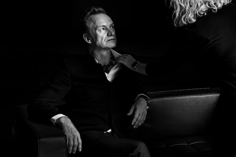 A black-and-white image of a man sitting on a couch, looking thoughtful. An unseen person stands nearby, adjusting his shirt collar. The scene is dramatic, with strong shadows and highlights.