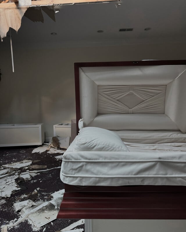 A partially open coffin sits in a room with debris scattered on the floor. The ceiling is damaged with large pieces hanging down, exposing insulation. The walls are bare and a few empty shelves are visible.