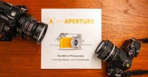 A book titled "A is for Aperture: The ABC's of Photography" by Todd Vorenkamp, illustrated by Jentana Shapiro, rests on a wooden surface. Two vintage cameras flank the book, one on the left and one on the right.