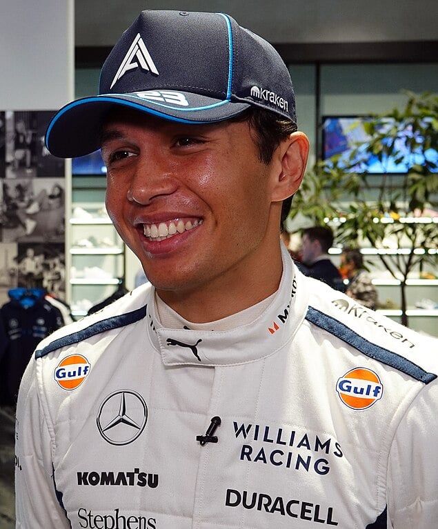 A person in a Williams Racing suit and cap is smiling. There are sponsor logos on the suit and cap, including Duracell, Mercedes, and Gulf. The background is blurred, with people and a plant visible.