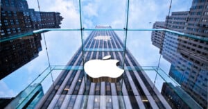 View looking up at a glass building with the Apple logo prominently displayed. Tall skyscrapers surround the structure under a partly cloudy sky. The architecture features reflective glass panels and a modern design.