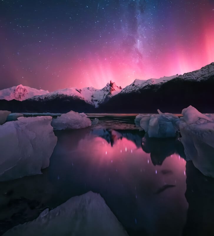 A stunning view of the Northern Lights illuminating the sky in pink and purple hues over a serene icy landscape with snow-covered mountains and large icebergs reflected in the calm water below. A starry night sky adds to the ethereal scene.