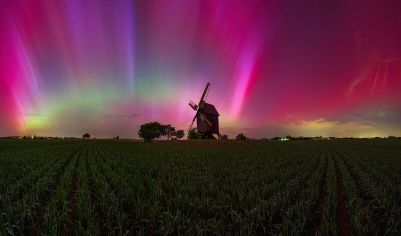 A vibrant aurora borealis displays shades of pink, purple, and green over a field with rows of small plants. A dark windmill stands silhouetted against the colorful sky. Bright lights are on the horizon under the starry night.