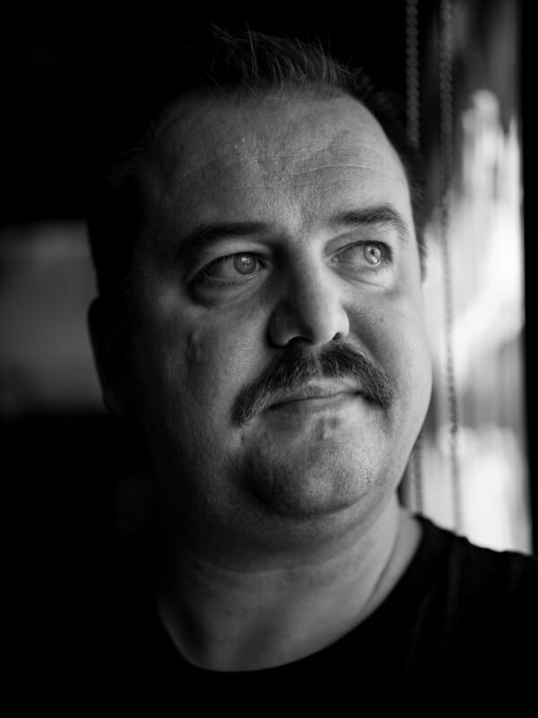 Black and white portrait of a man with a mustache, looking thoughtfully out of a window, with light falling on his face from the side. The expression is contemplative and the background is blurred.