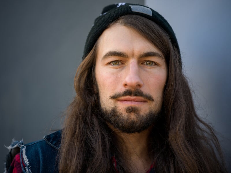 A person with long brown hair and a beard wearing a black beanie and a denim jacket. They are standing against a gray background, looking directly at the camera with a neutral expression.