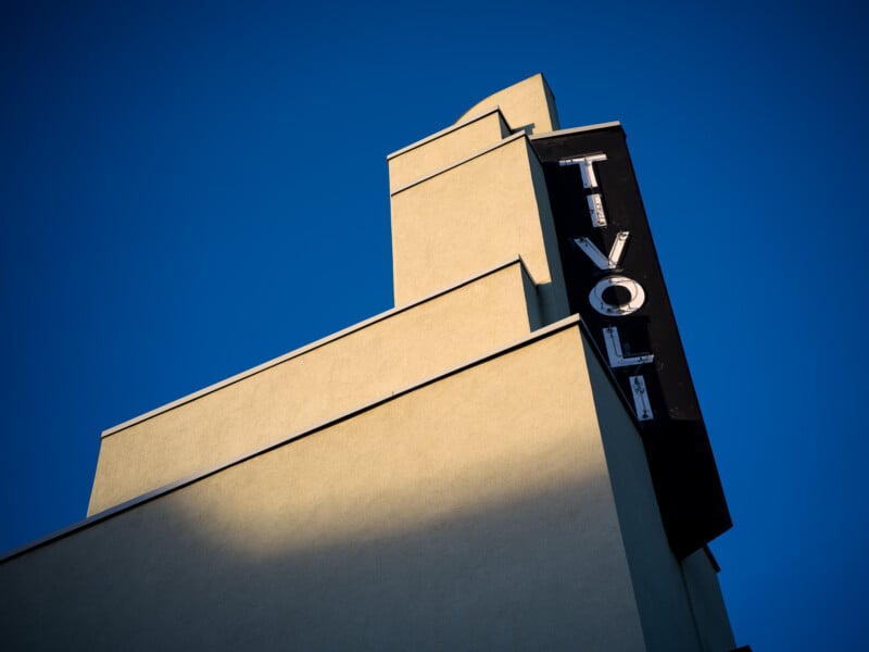 A building with a minimalist architectural design features a vertical sign that reads "TIVOLI." The structure is bathed in warm sunlight against a clear blue sky, highlighting its geometric shapes and modern style.