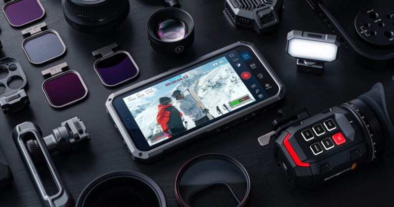 A flat lay of various camera equipment and accessories surrounding a smartphone displaying a snowy mountain scene with people. The setup includes lenses, filters, a camera module, and other electronic gear on a dark surface.