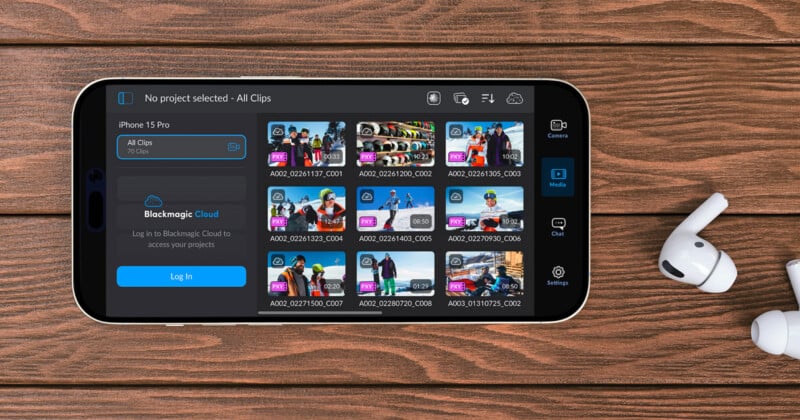 A smartphone laid on a wooden surface displays a media app interface with various video clips. Next to it are wireless earbuds. The screen shows video thumbnails and Blackmagic Cloud login options.