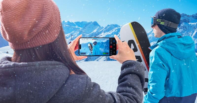 A person in a brown beanie takes a photo with a smartphone of a snowboarder on a snowy mountain. The snowboarder, wearing a blue jacket and holding a board, looks at the snowy peaks under a clear blue sky.