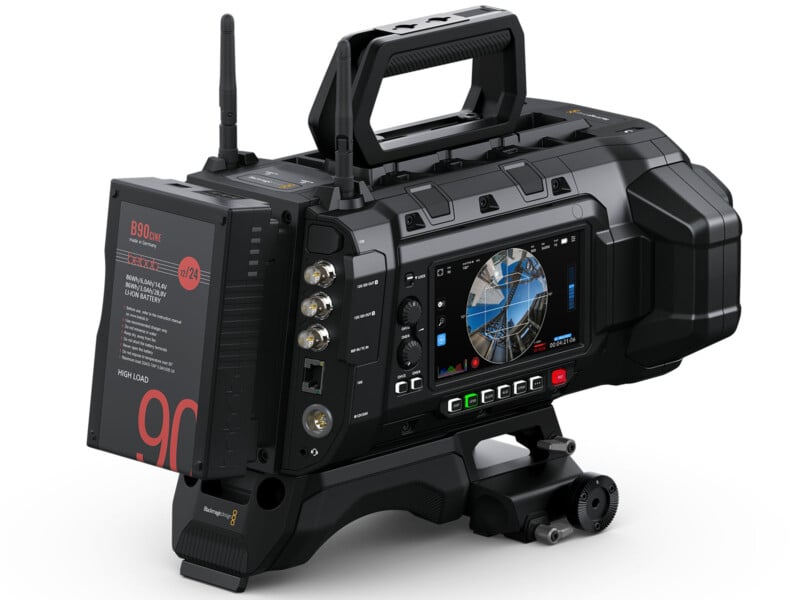 A professional black cinema camera with various buttons and a color display screen. It features a handle on top and a mounting attachment at the bottom.