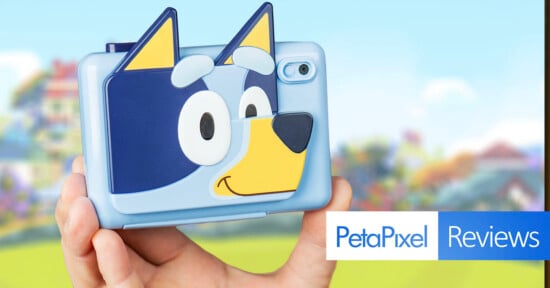 A person holds a blue camera with a cartoon dog face design on the back. The background is a blurred colorful outdoor scene. The bottom right corner shows "PetaPixel Reviews" text.