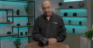 A person in a black Canon shirt stands behind a wooden table with a Canon camera on it. Shelves in the background display various camera lenses and small plants. The room is softly lit with blue ambient lighting.