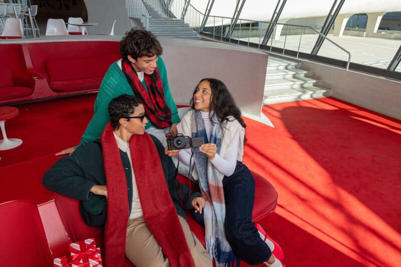 Three people in a modern lounge with red seating are sharing a camera and smiling. Two are sitting, wearing scarves and glasses, while the third is standing. Large windows in the background let in natural light.