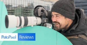 A person in winter clothing smiles while holding a camera with a large telephoto lens. They are positioned behind a green sculpture. The image also features the "PetaPixel Reviews" logo in the corner.