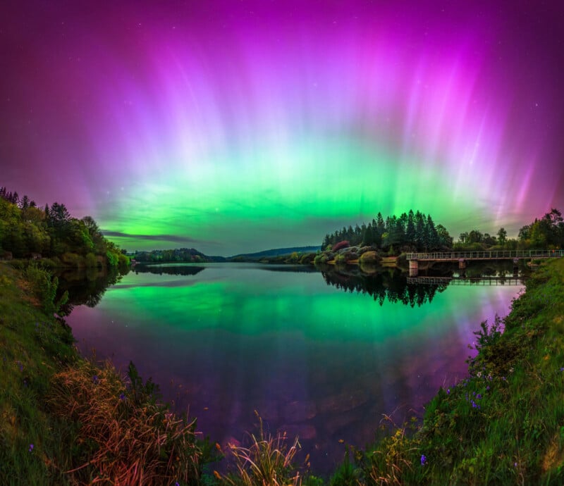 A vivid display of the Northern Lights with radiant green and purple hues filling the sky, reflecting on a calm, lake surrounded by trees and a bridge. The scene is serene and ethereal, showcasing nature's beauty.