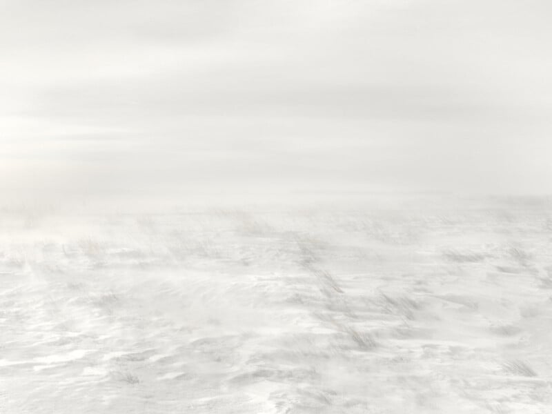 A vast, snowy landscape under a cloudy sky, with subtle, blurred details suggesting wind-swept snow and sparse vegetation barely visible through the white expanse. The atmosphere conveys a sense of cold and desolation.