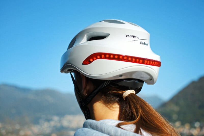 Person wearing a white bike helmet with LED light on the back, facing away toward a scenic view of mountains and a blue sky.