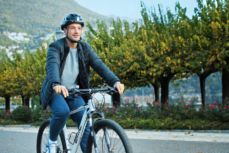 A person wearing a helmet and a dark jacket rides a bicycle on a path lined with trees. The background features green hills under a partly cloudy sky.