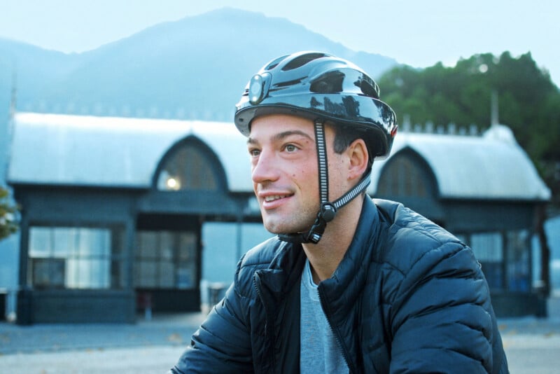 A person wearing a bike helmet and a jacket is smiling outdoors. In the background, there is a large building with arched windows and a mountain under a cloudy sky.