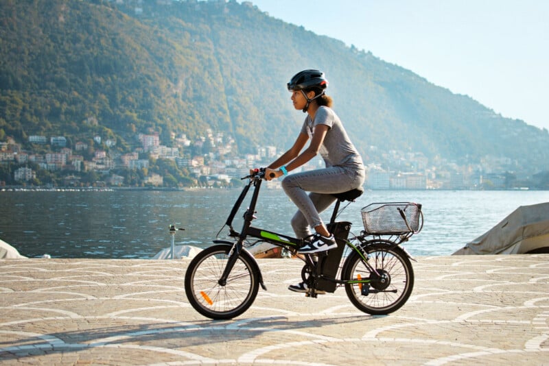 A person in a gray outfit and helmet rides a black bicycle with a basket along a waterfront promenade. A scenic hillside with buildings and trees is visible in the background under a clear blue sky.