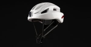 A sleek white helmet with black interior padding and adjustable chin straps is suspended against a dark background. The helmet features ventilation openings and a small circular opening at the front.