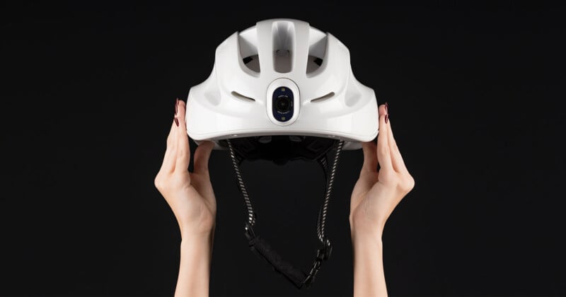 Hands holding a white bicycle helmet with a camera on the front, set against a black background.