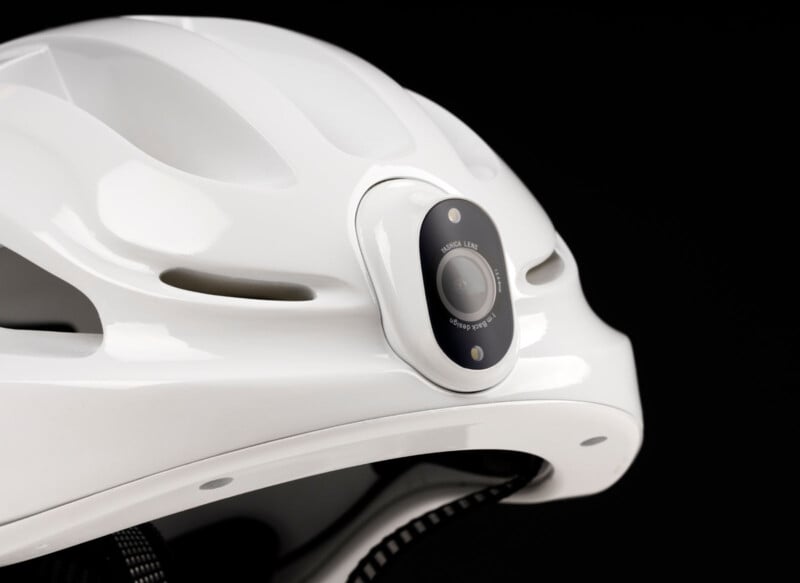 A close-up of a white helmet featuring a built-in black camera on the front. The helmet has a smooth, glossy surface and ventilation slots, set against a black background.