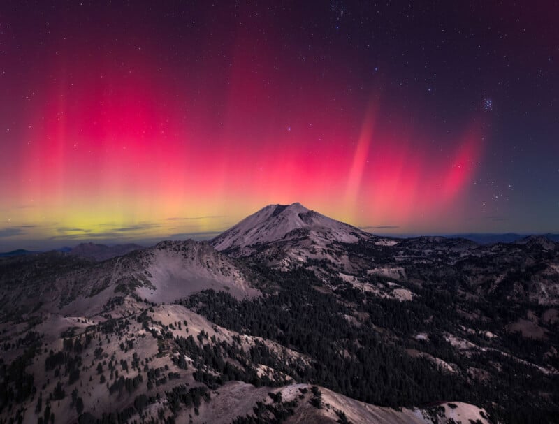 A majestic mountain landscape under a starry night sky with vibrant pink and red auroras. The mountain peaks and rugged terrain are illuminated by the colorful northern lights, creating a stunning natural scene.