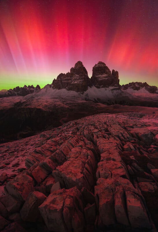 A stunning mountain landscape under a vibrant red aurora. Jagged rocky foreground leads to tall peaks silhouetted against the colorful sky. The night sky is filled with stars, adding to the enchanting scene.