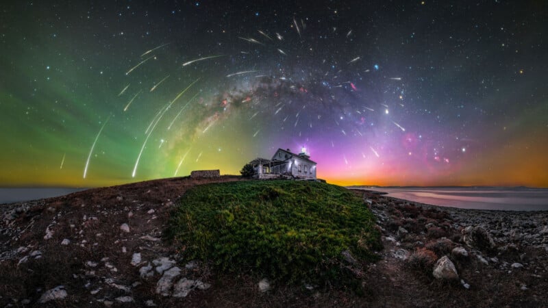 A rustic house sits atop a hill surrounded by greenery, under a starry night sky. The sky displays a colorful aurora, meteor streaks, and the Milky Way, casting a magical glow over the landscape.