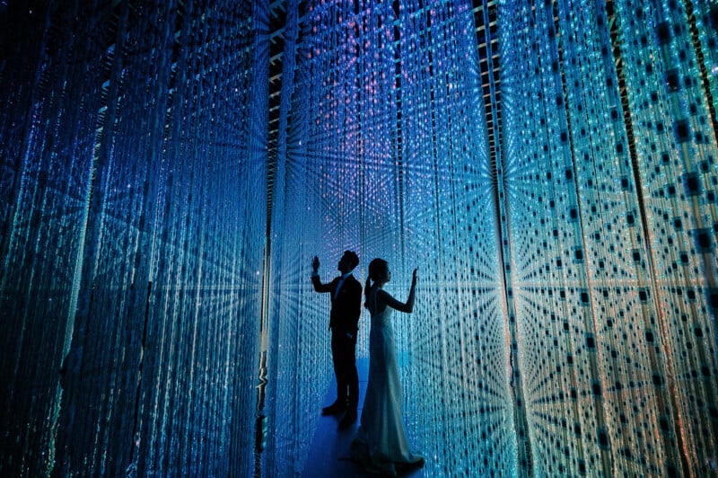 Two silhouetted figures stand in a mesmerizing light installation with blue lights and patterns surrounding them. The scene has a futuristic and dreamy atmosphere.