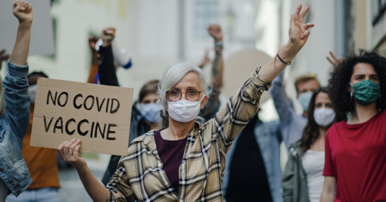 A group of people are protesting outdoors. A woman in the foreground with glasses and a mask holds up a sign reading "NO COVID VACCINE." Others in the background also have raised fists and are wearing masks.