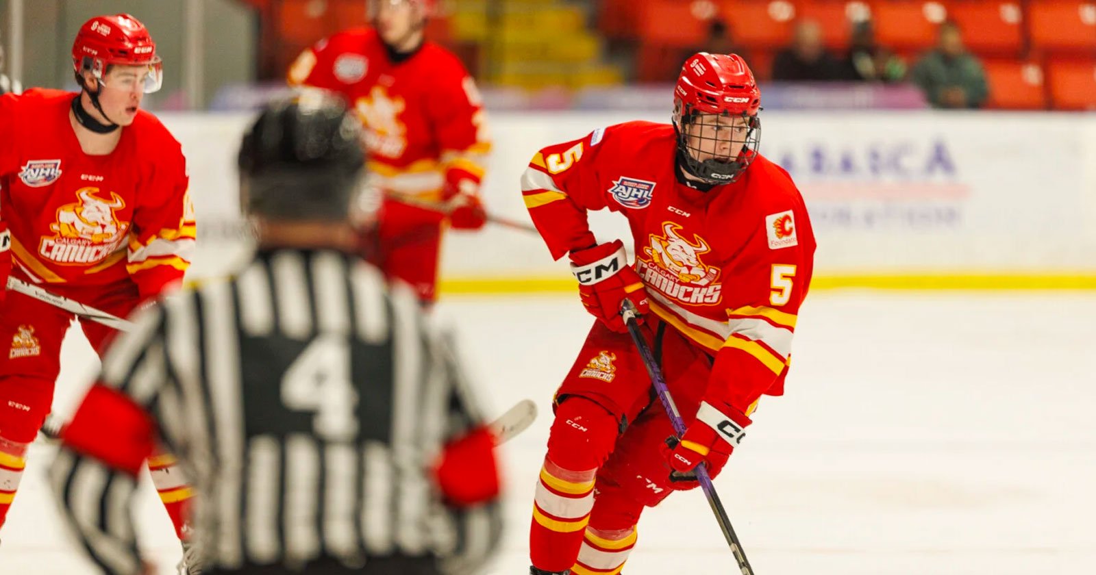 Hockey players in red uniforms compete on the ice, with one player in focus wearing a helmet and jersey number 5. A referee, blurred in the foreground, wears a black and white striped shirt with the number 4. The background shows empty arena seats.