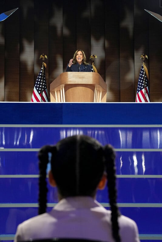 A woman speaks passionately at a podium on a stage adorned with American flags. The focus is on a young girl with braided hair watching the speaker intently from the audience.