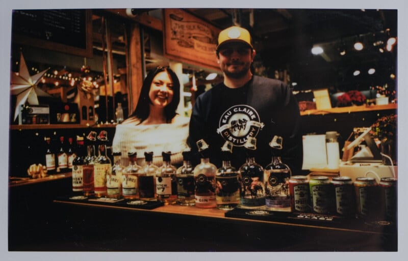 A woman and a man stand behind a bar, displaying various bottles of liquor and canned drinks. The bar has a rustic interior with dim lighting, creating a cozy atmosphere.