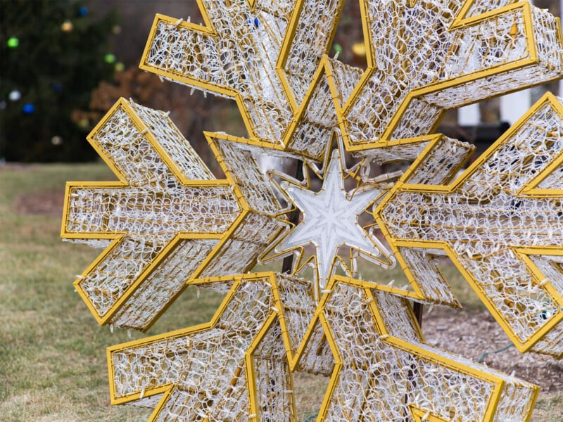 Close-up of a large decorative metal star with intricate wire mesh and lights, displayed outdoors on grass. The star features a golden frame with multiple pointed geometric layers and a smaller star at its center.