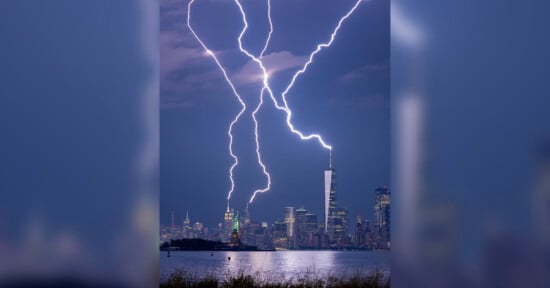 A dramatic scene of multiple lightning bolts striking behind a city skyline at night. Prominent buildings are visible, and the sky is dark and cloudy, with bright lightning illuminating the scene.