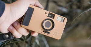 A person holds a vintage-style point-and-shoot camera with a tan body and black accents. The camera has a large lens, flash, and the text "Film Never Die" on the front. The background is blurred foliage.