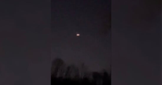 A dark sky with a faint, glowing object resembling a circular light is visible in the center. Silhouetted trees are at the bottom of the image, suggesting a nighttime scene.