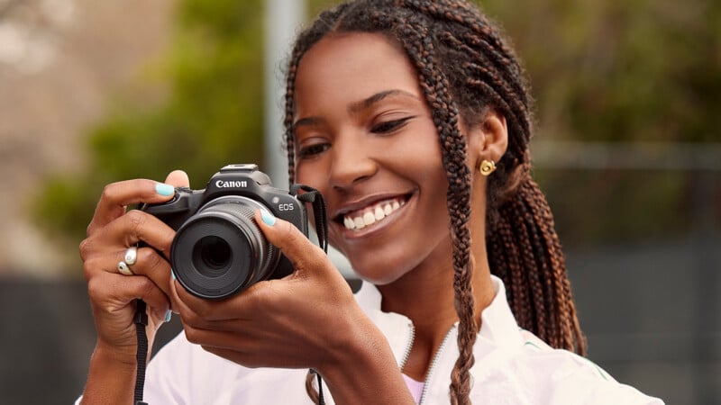 A person with braided hair is smiling while holding a Canon EOS camera, looking at the screen. They are outdoors with blurred greenery in the background, wearing a white jacket and small hoop earrings.