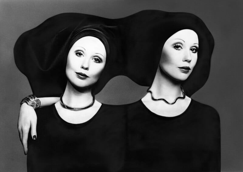 Black and white image of two women wearing dark clothing and head coverings, their faces leaning towards each other. One has her arm draped over the other's shoulder. Both wear statement necklaces and are gazing forward.