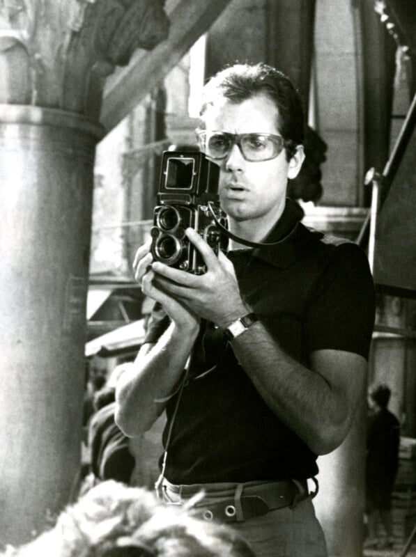 A person wearing sunglasses holds a vintage camera, focusing intently. They stand outdoors near architectural columns in a black-and-white photograph, suggesting a setting of meticulous photography or filming.