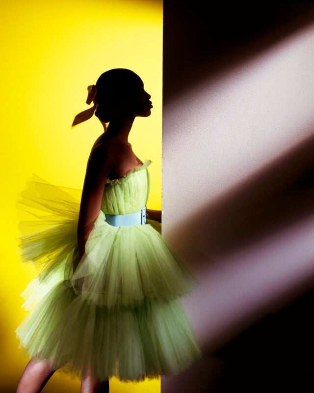 Silhouette of a person in a green tulle dress with a ribbon in their hair, standing against a vibrant yellow background. They are partially illuminated by light streaming from the right, creating a dramatic contrast.