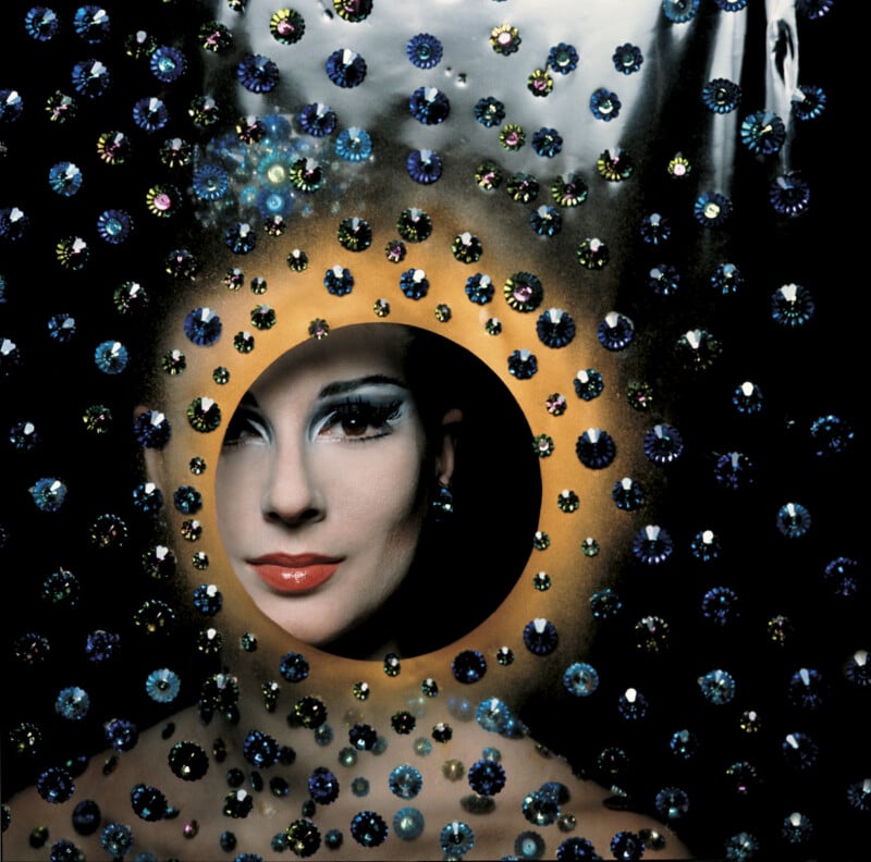 A woman with dramatic makeup peers through a circular opening surrounded by an array of sparkling, colorful gems on a dark background. The gems create an artistic, almost celestial pattern around her.