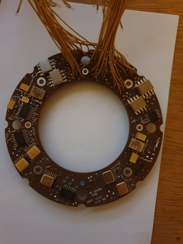 A circular printed circuit board with multiple electronic components and numerous yellow wires extending from it, placed on a piece of white paper on a wooden surface.