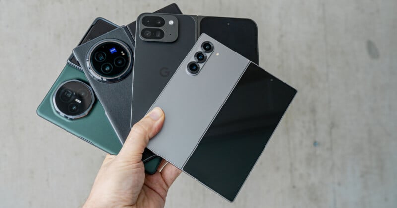 A hand holds five different smartphones fanned out, showcasing various designs and camera styles. The phones vary in size, color, and brand, with prominent camera modules visible. The background is a plain, light-colored surface.